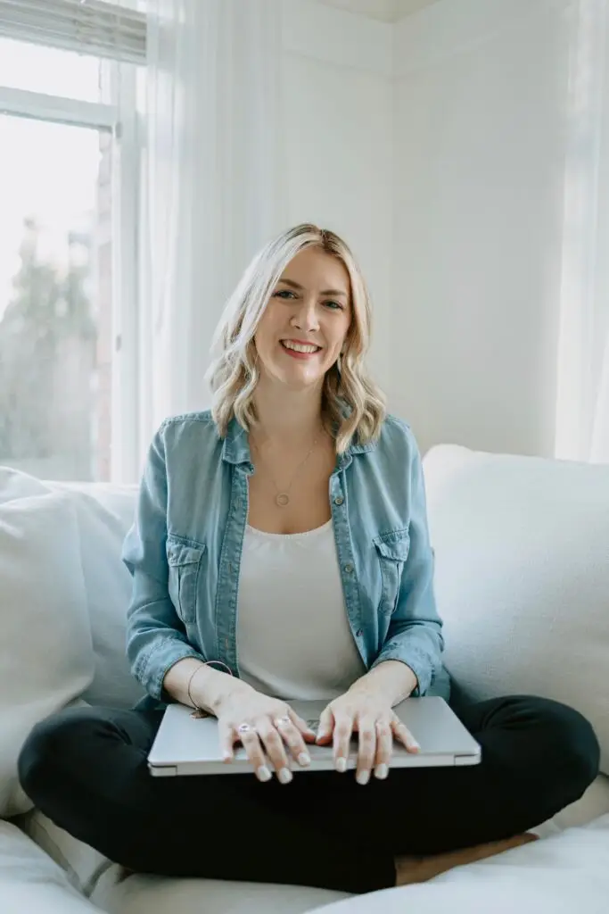Ashley is sitting cross-legged on a white sofa with a closed laptop on her lap.