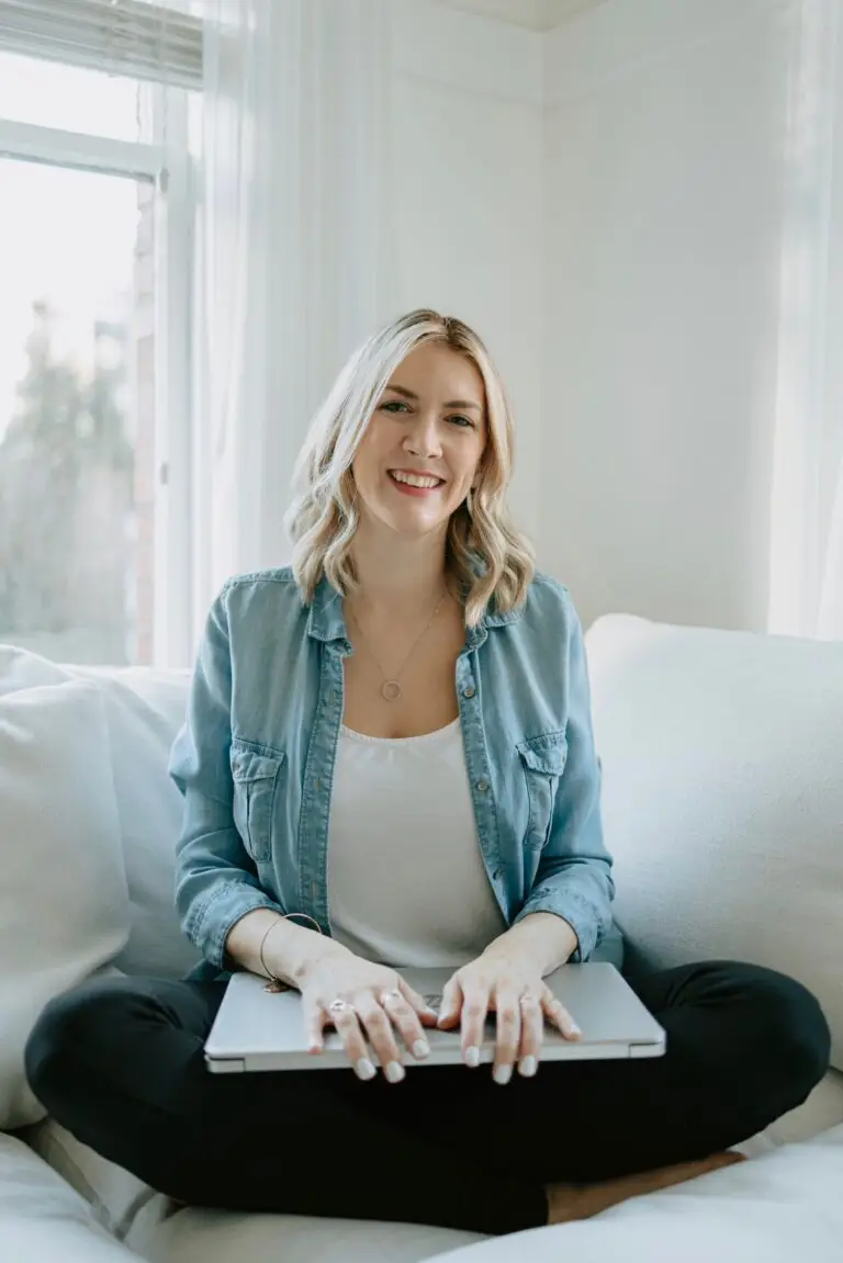 Ashley is sitting cross-legged on a white sofa with a closed laptop on her lap.